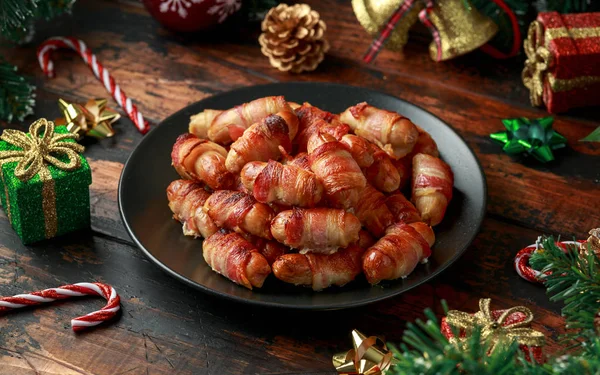 Christmas Pigs in blankets, sausages wrapped in bacon with decoration, gifts, green tree branch on wooden rustic table — Stock Photo, Image