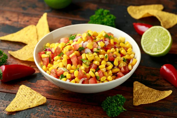 Mexican Corn Salsa in white bowl with lime and tortilla chips on wooden table. Healthy food