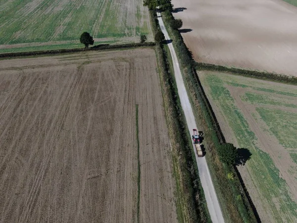 An aerial view of a tractor and trailer on a long straight road with hedgerow and ploughed fields either side — Stock Photo, Image