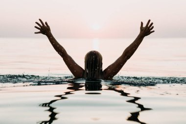 Denizde bir kadın, gün batımında kolları suyun üstünde bakıyor.