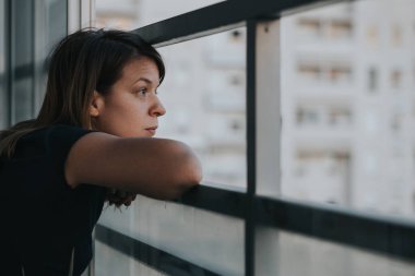 Genç üzgün kadın bir apartmanın balkonundan dışarı bakıyor.
