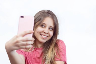 Pembe tişörtlü, selfie çeken beyaz, mutlu bir kızın portresi. İzole edilmiş beyaz arka plan. Gerçekten samimi duygular. Güzel bir gülüşü olan genç bir kadın. Doğal renkte dişler bile.