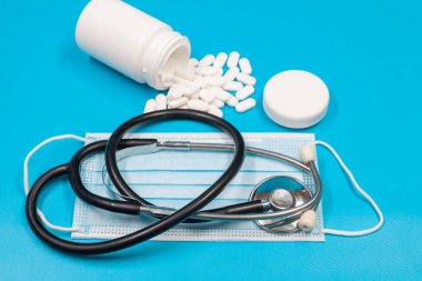 Stetoskop, cerrahi maske. Beyaz haplar mavi arka planda beyaz konteynırdan dökülüyor. Seçici odaklanma. Kapat..