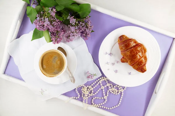 Breakfast coffee and croissant served on lavender bed tray with a bunch of lilac, napkin, strand of pearls.