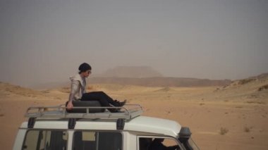 Arabanın çatısındaki adam çölde seyahat ediyor.