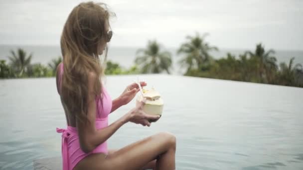 여자는 수영장에서 휴식을 취하고 코코넛 주스를 마시고 아름다운 몸매의 수영복을 — 비디오