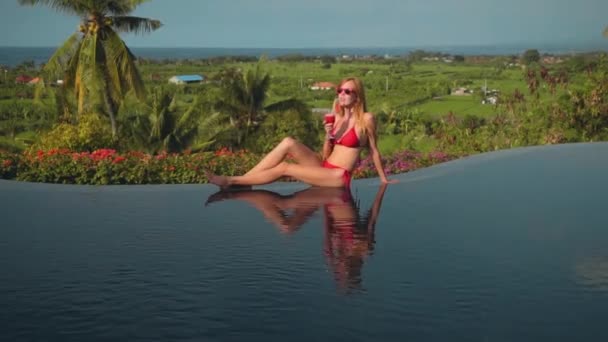 Junge Frau im roten Bikini trinkt Saft am Rande des Infinity-Pools, Meerblick — Stockvideo