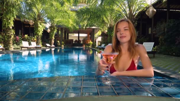 Девушка пьет коктейль в баре у бассейна под пышными пальмами — стоковое видео