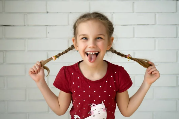 Adorable chica divertida sosteniendo coletas de pelo en una pared de ladrillo. Pequeña linda chica con el pelo largo y rubio al aire libre jugando, mostrando la lengua —  Fotos de Stock