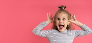 Bun saçlı, gri çizgili bluz giyen sarışın çocuk. Surat yapıyor, pembe stüdyo geçmişine karşı poz veriyor.