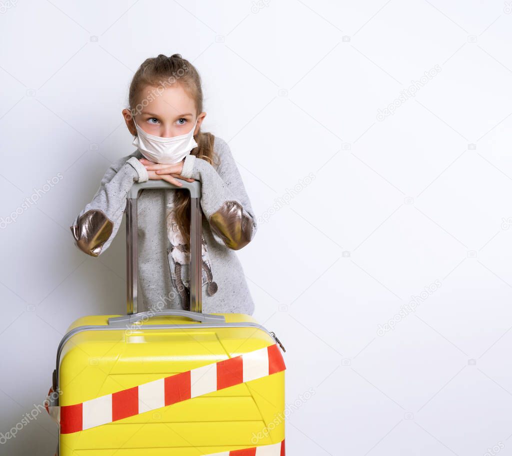 Blonde kid in gray jumper, medical mask. Posing isolated on white. She leaning on yellow suitcase tied white and red warning tape