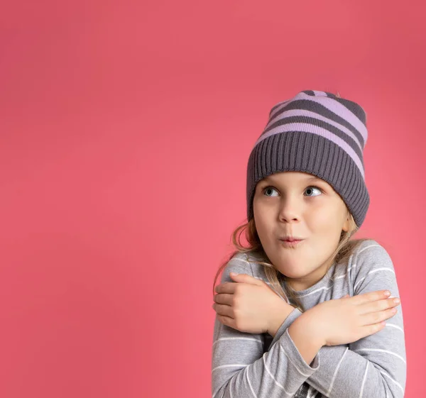 Ragazzina bionda con camicetta e cappello a righe grigie. Si abbraccia, sembra congelata, posa sullo sfondo dello studio rosa — Foto Stock