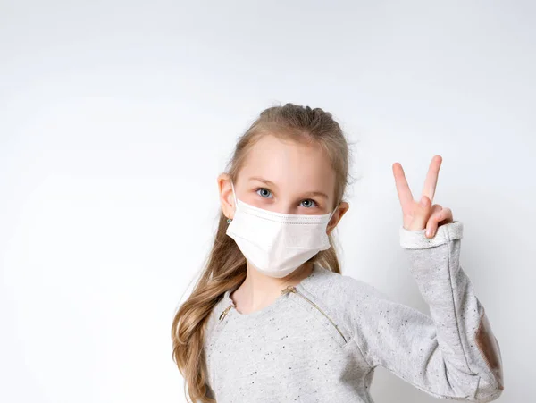 Gri kazaklı ve tıbbi maskeli küçük sarışın kız, beyaz üzerinde yalnız poz veriyor. Zafer eli gösteriyor. Coronavirus. Karantina