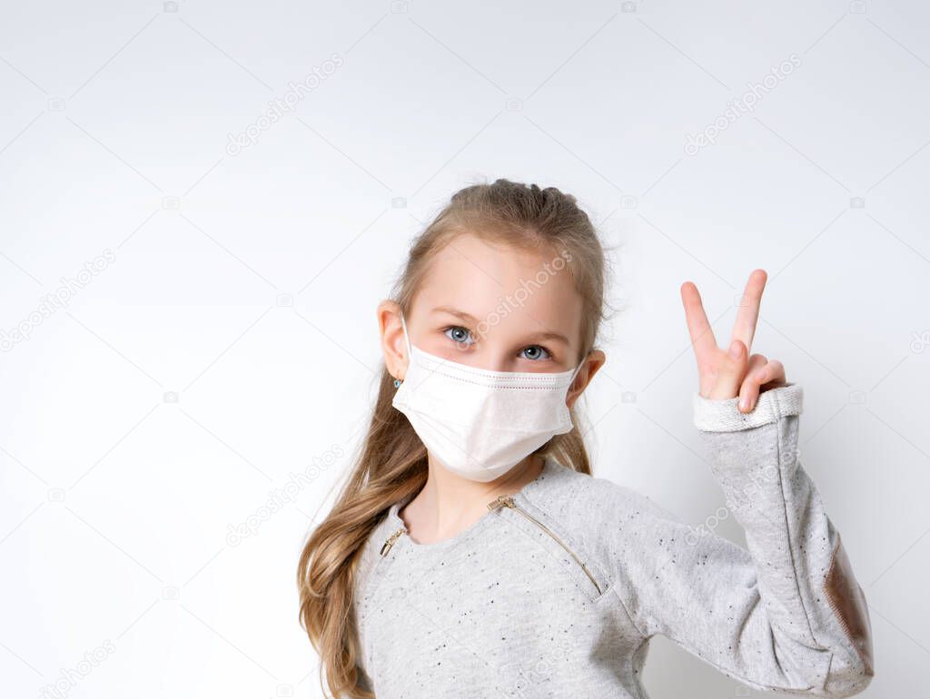 Blonde little girl in gray jumper and medical mask, posing isolated on white. She showing victory hand. Coronavirus. Quarantine