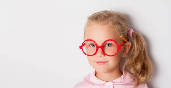 Niño rubio con cola de caballo, en gafas enmarcadas rojas, blusa rosa. Sonriendo, posando con lápiz amarillo detrás de la oreja. Aislado sobre blanco —  Fotos de Stock