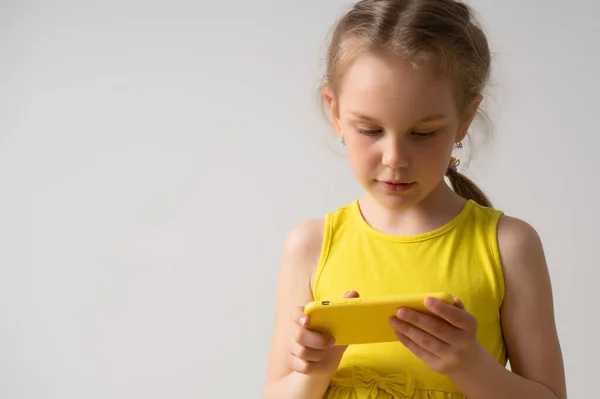 La niña seria concentrada en vestido amarillo brillante está involucrada en jugar juegos en línea en su teléfono móvil. Retrato de cerca aislado en blanco — Foto de Stock