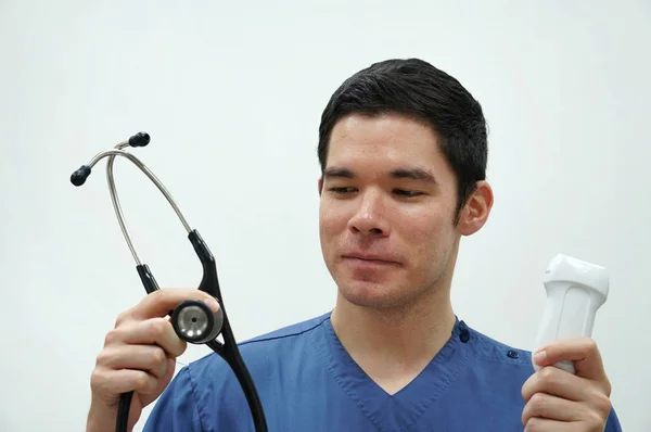 Un jeune homme beau en uniforme de gommage bleu tenant stéthoscope et sonde à ultrasons sur le fond blanc — Photo