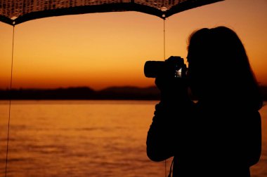 A silhouette of woman holding a camera taking a photo of river view from the boat at the sunset                       