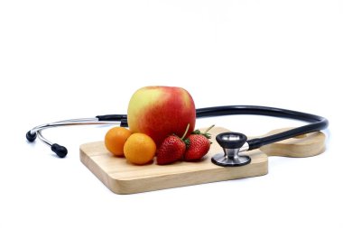 Siyah steteskop, kırmızı elma, küçük portakallar ve beyaz arka plandaki tahta kesme tahtasının üzerinde çilekler.