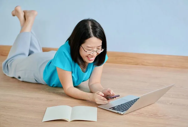 Een jonge Aziatische vrouw met blauwe blouse en bril op een houten vloer met behulp van een smartphone met computer en boek op een vloer — Stockfoto