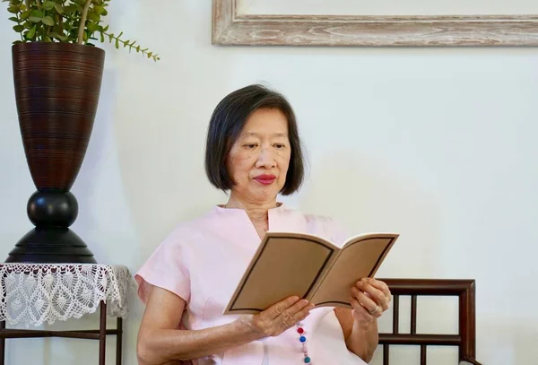 An old asian woman in pink blouse sitting on a chair reading in a house