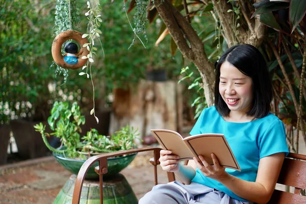 Eine junge asiatische Frau in blauer Bluse liest ein Buch und sitzt auf einer Holzbank im Garten — Stockfoto
