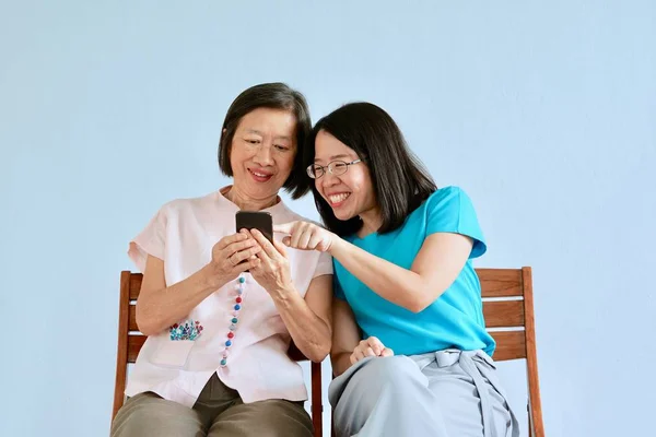 Uma filha asiática em blusa azul ensinando sua mãe asiática para usar telefone inteligente no quarto azul — Fotografia de Stock