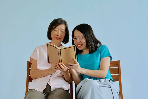 An asian old mother in pink blouse sitting on a chair reading a book with a young asian woman in blue blouse sitting on a chair