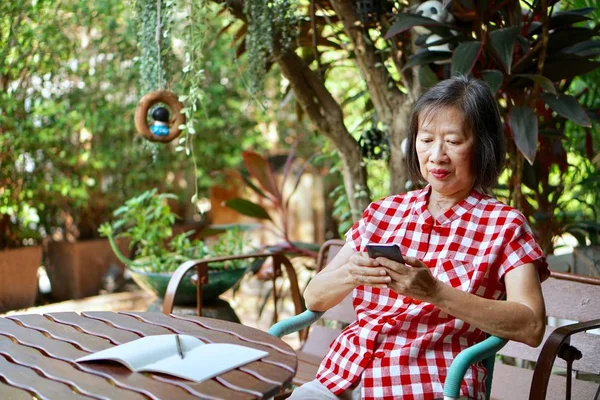 stock image An old asian woman with red blouse siiting on the bench using black smart phone in the garden