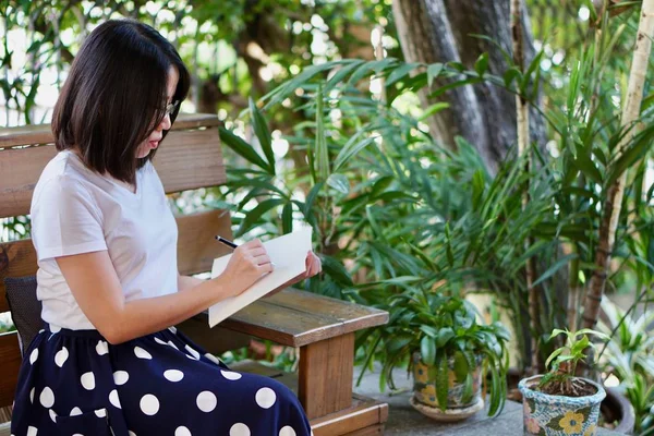 Eine junge schöne asiatische Frau mit Brille, weißem T-Shirt und blauem Rock sitzt auf der Bank und schreibt ein Tagebuch im Garten — Stockfoto