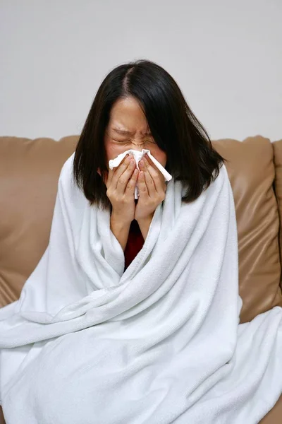 Une jeune femme asiatique assise sur le canapé brun couvrant avec couverture blanche ayant froid commun — Photo