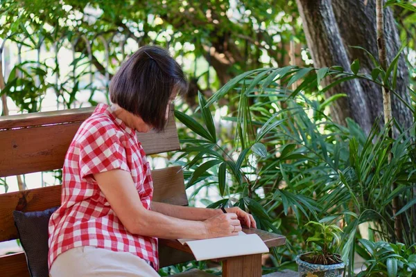 Eine alte asiatische Frau in roter Bluse schreibt Tagebuch im Garten — Stockfoto