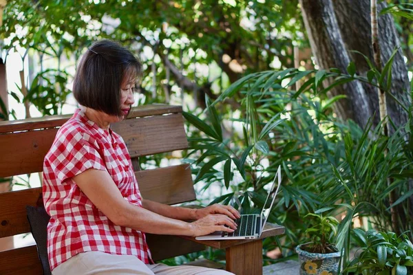 An old asian woman with red blouse sitting on the bench playing computer in the garden