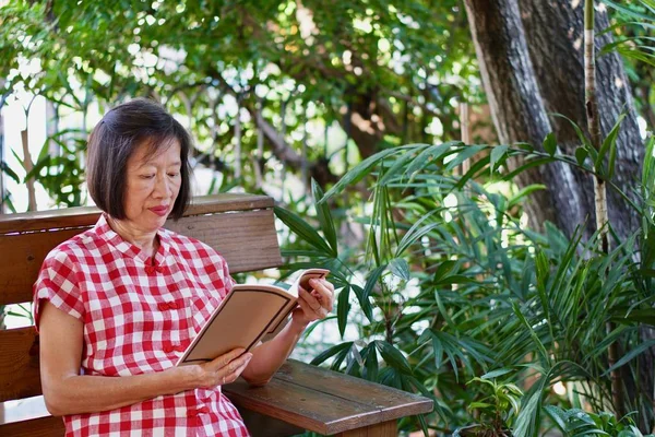 Eine alte asiatische Frau in roter Bluse liest im Garten ein Buch — Stockfoto