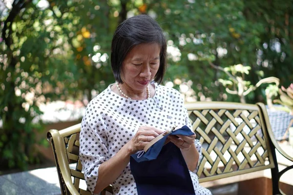 Una vieja asiática sentada en el banco bordando pañuelo en el jardín — Foto de Stock