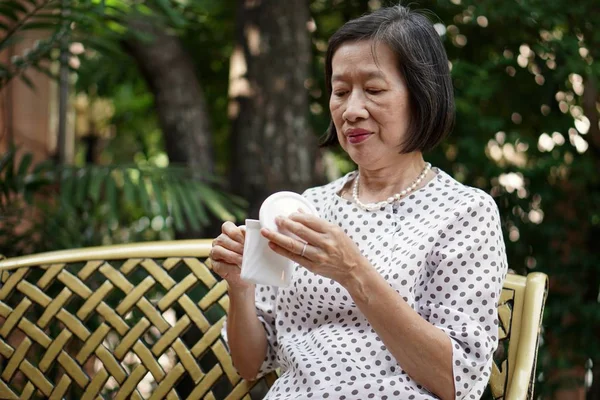 Una anciana asiática con lunares marrones en blusa blanca sentada en el banco bebiendo té en el jardín — Foto de Stock