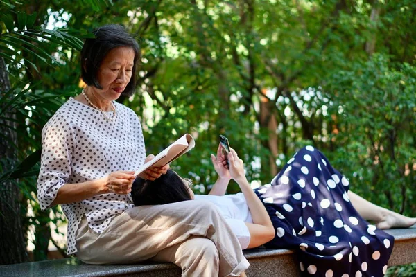 Una madre con una blusa blanca de lunares leyendo un libro sentado en un banco con una hija con lunares blancos en falda azul usando un teléfono inteligente tumbado en el regazo de la madre — Foto de Stock