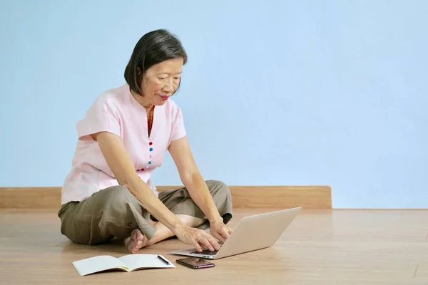 Uma mulher asiática velha na blusa rosa com cara do sorriso usando o computador, sentada no assoalho de madeira em uma sala azul — Fotografia de Stock