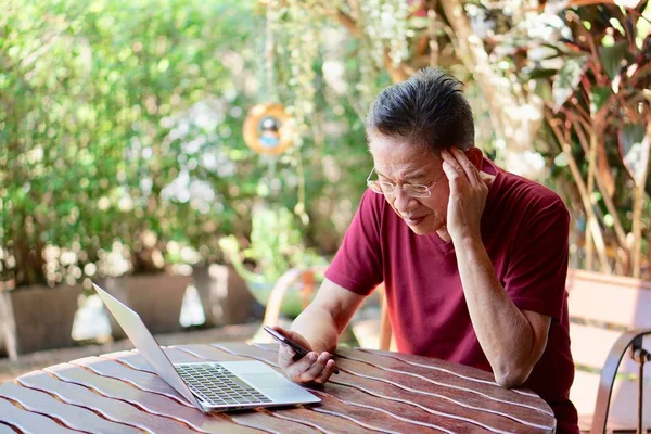 An old asian man in red t shirt and eyeglasses using smart phone and getting headache, with a computer on the table, in the garden