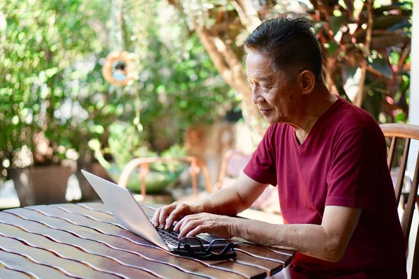 An old asian man in red t shirt sitting on a wooden chair using computer in a garden