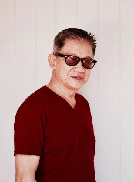An old asian man in red t shirt and sunglasses standing in front of white wall