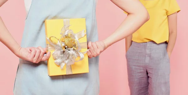 Una mujer en jean azul escondiendo una caja amarilla detrás de su espalda y una mujer en blusa amarilla esperando un regalo, fondo rosa — Foto de Stock