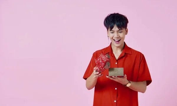 A young asian man with happy face and red shirt openning a present box, standing on the pink background