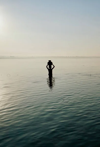 Silueta de un niño sosteniendo un tarro de bambú sobre la cabeza, de pie en el agua al amanecer —  Fotos de Stock