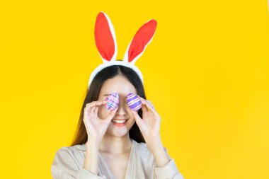 Gülümseyen yüzlü, tavşan kulağı takan, sarı arka planda çok renkli paskalya yumurtaları tutan genç bir Asyalı kadın.