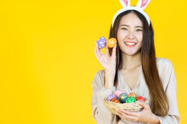 Gülümseyen yüzlü, tavşan kulağı takan, sarı arka planda çok renkli paskalya yumurtaları tutan genç bir Asyalı kadın.