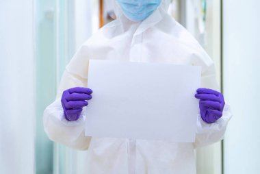 Beyaz takım elbise maskesi ve eldivenler de dahil olmak üzere kişisel koruyucu ekipmanlı bir doktor elinde kelime doldurmak için boş bir kağıt tutuyor.
