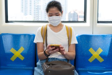 Yüz maskesi takan genç Asyalı kadın, akıllı telefon kullanıyor ve gökyüzü treninde oturuyor. Covid-19 ya da Coronavirus salgını sırasında diğer insanlarla arasına sosyal mesafe koyuyor.