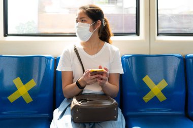 Yüz maskesi takan genç Asyalı kadın, akıllı telefon kullanıyor ve gökyüzü treninde oturuyor. Covid-19 ya da Coronavirus salgını sırasında diğer insanlarla arasına sosyal mesafe koyuyor.