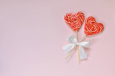 Pembe bir arka planda, sağ tarafında beyaz bir fiyonk olan tahta bir çubukta kalbi olan iki parlak lolipop. Kartpostal Boş Boşluk Üst Görünümü Düzeni. Minimalizm. Sevgililer Günü.